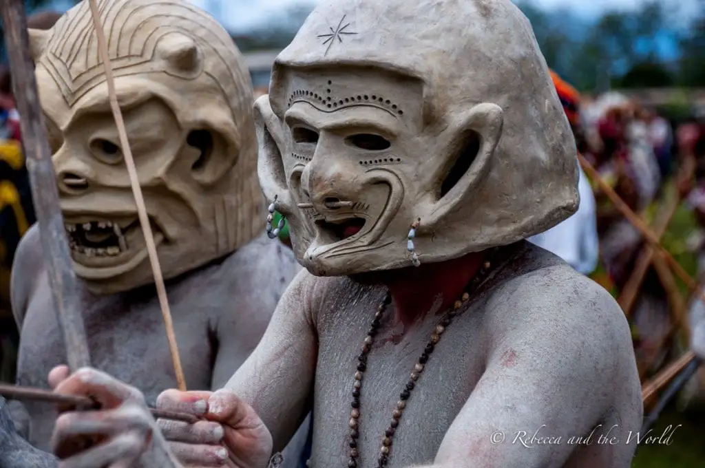 Two individuals wearing traditional mud masks with carved designs at a Papua New Guinea festival. The masks feature expressive faces and are accessorized with simple necklaces, enhancing their tribal look. The famous Asaro Mudmen can be seen at the Hagen Show and Goroka Show in Papua New Guinea.