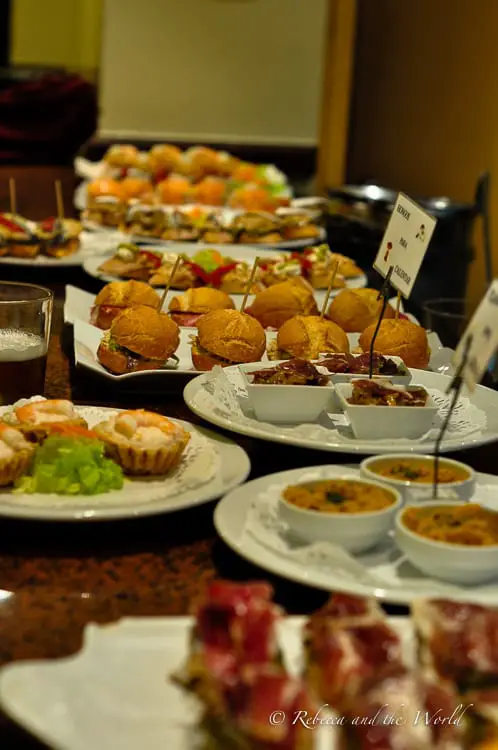 A close-up of an assortment of pintxos, traditional Basque small snacks, arranged on white plates on a bar counter. They appear freshly prepared, with a variety of toppings, including meats and vegetables, served on slices of bread.