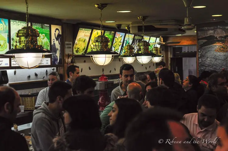 A busy and vibrant bar with patrons gathered around the counter, engaged in conversation. The atmosphere seems lively and social, with illuminated menu boards and vintage style hanging lamps above.