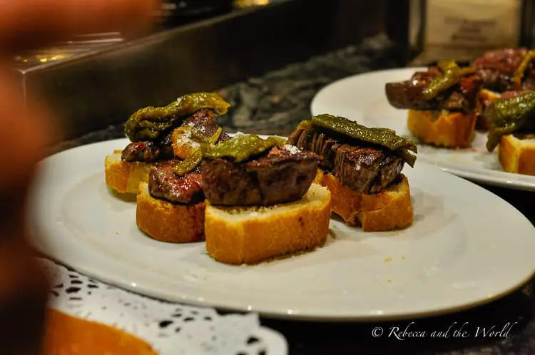 A close view of a plate of pintxos featuring small slices of steak topped with green peppers, served on crusty bread. The arrangement is simple and appetising, highlighting the pintxos as a culinary attraction.