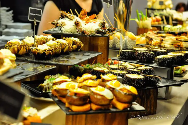A close-up of an assortment of pintxos, traditional Basque small snacks, arranged on white plates on a bar counter. They appear freshly prepared, with a variety of toppings, including meats and vegetables, served on slices of bread.