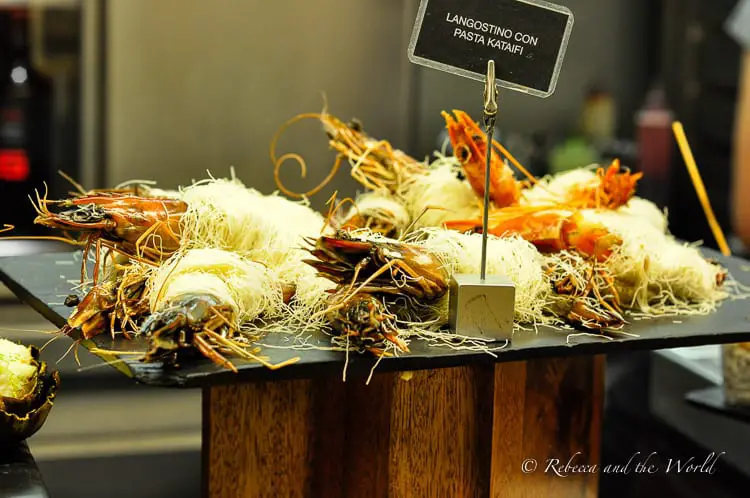 An elaborate display of large prawns resting on a bed of thin, straw-like noodles. A small sign identifies the dish as "Langostino con pasta kataifi". The presentation is creative and visually striking, emphasising the gourmet aspect of the pintxos tour experience.