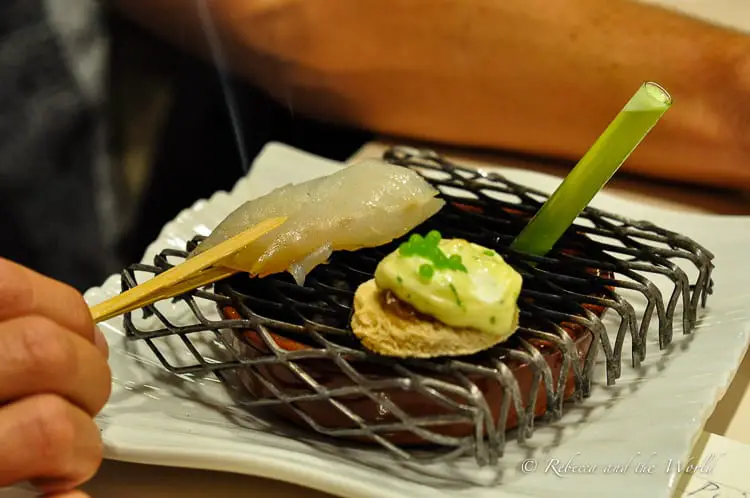 An artistic presentation of a single pintxo consisting of a skewered piece of seafood on a small round of bread, accented with a green sauce and a garnish, displayed on a wire stand with a minimalist design.