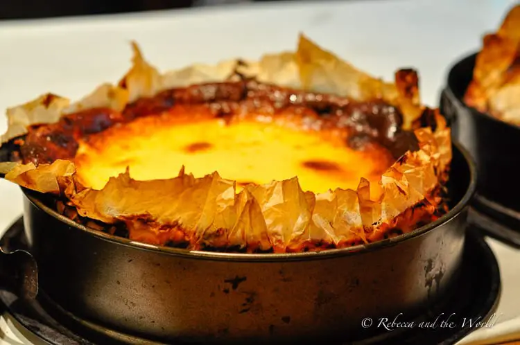 A rustic baked Basque cheesecake, displayed in a round baking tin. The top is flaky, suggesting a light, crispy texture, and it is served directly from the oven.