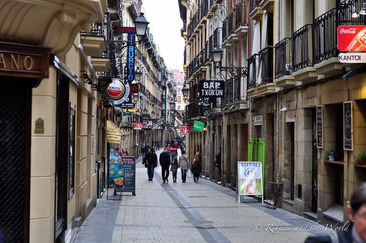 A narrow cobblestone street lined with classic European buildings featuring iron balconies. Various signs and flags are displayed above the street, indicating bars and other businesses. Pedestrians are walking down the sidewalk, creating a sense of everyday life in this urban setting. This is San Sebastian, home of pintxos!