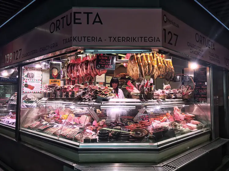 A well-lit charcuterie stall displaying an array of Spanish cured meats and sausages, highlighting the importance of gastronomy in Bilbao. The La Ribera Market is one of the best places to visit in Bilbao, Spain.