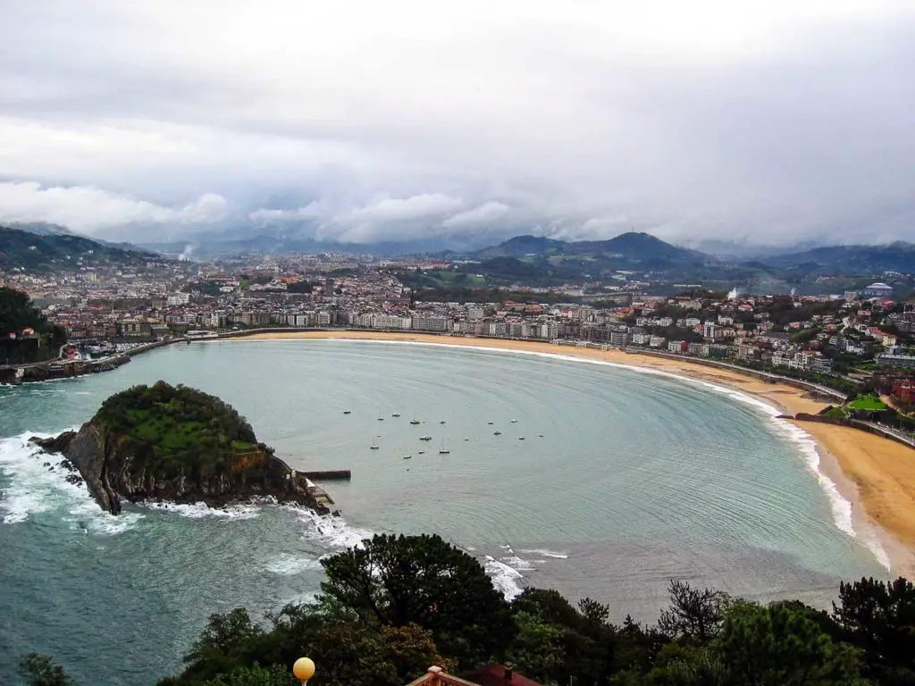 An expansive view of San Sebastian's bay, featuring the crescent-shaped La Concha Beach with calm waters and boats. A lush green island is at the bay's entrance, and the city's buildings frame the shore against a backdrop of low clouds and mountains.