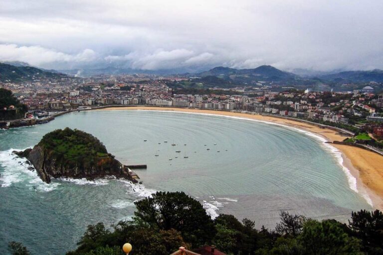 An expansive view of San Sebastian's bay, featuring the crescent-shaped La Concha Beach with calm waters and boats. A lush green island is at the bay's entrance, and the city's buildings frame the shore against a backdrop of low clouds and mountains.