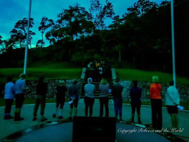 A group of people standing in a circle at dusk, participating in a Kokoda Track ceremony. They are facing the Isurava Memorial with a person speaking at the front, surrounded by flagpoles and trees.