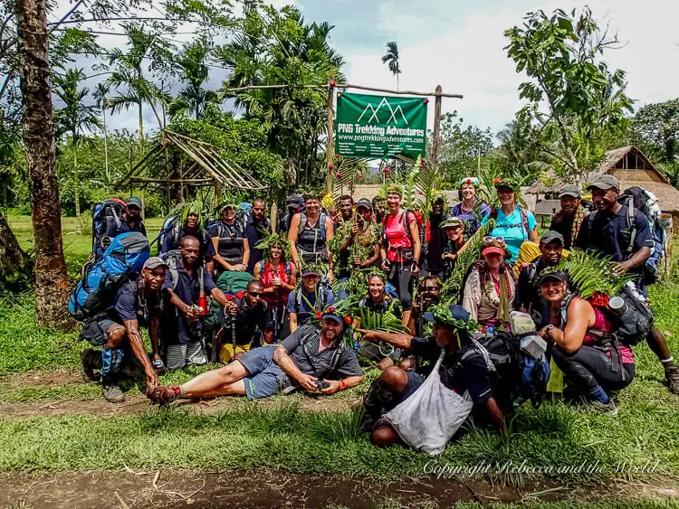 A large group of hikers and locals posing together for a group photo in front of a sign for "PNG Trekking Adventures." The group is smiling, holding trekking poles, and some are adorned with leafy crowns, indicating a celebratory moment.