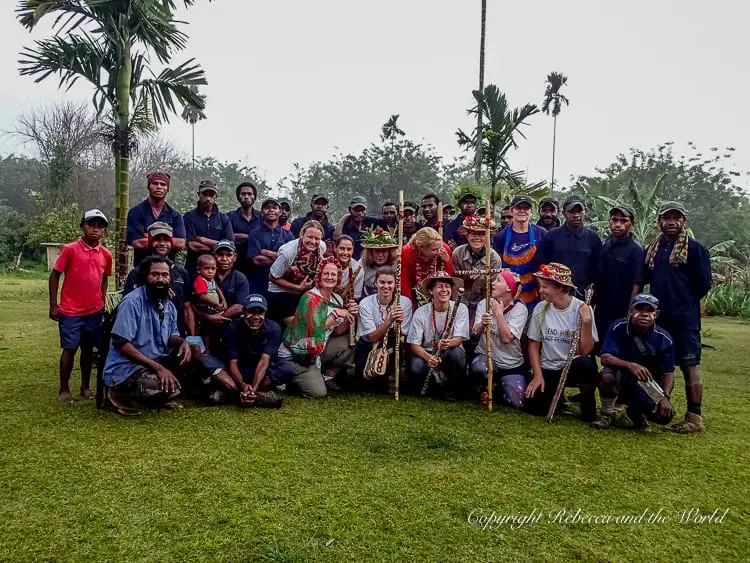 A large group of trekkers and local guides posing together for a group photo on a grassy area. The group includes men, women, and children, and some are wearing traditional attire, emphasizing the camaraderie and cultural exchange during the trek. This is our entire group of hikers, porters and guides on the Kokoda Track!