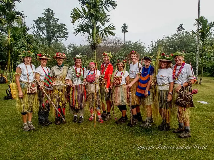 A group of trekkers wearing traditional grass skirts, flower crowns, and carrying walking sticks, posing together in a line on a grassy area. The background features tall palm trees and lush greenery, highlighting the celebratory and cultural aspect of the trek. This is the final day of our Kokoda Track experience.