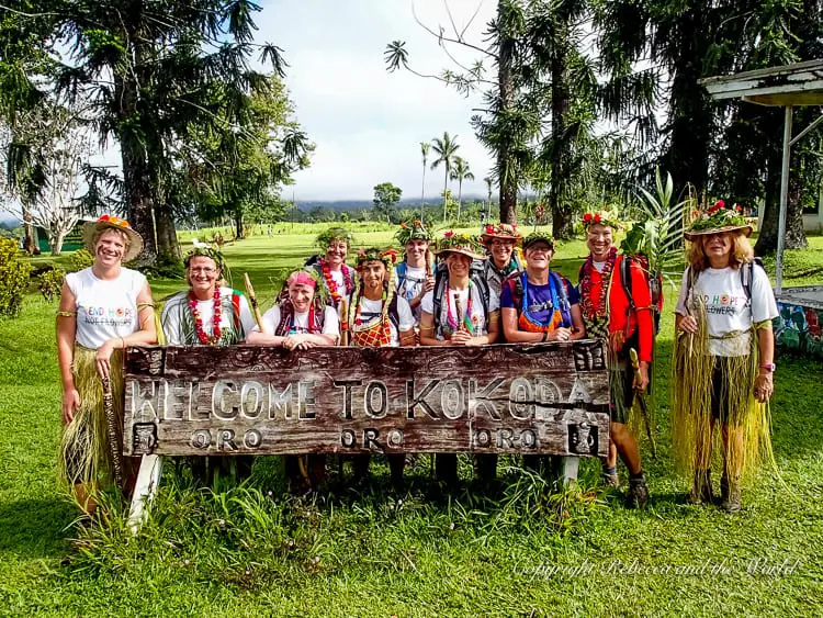 A group of trekkers in traditional grass skirts and flower crowns posing behind a large wooden sign that reads "Welcome to Kokoda." The background features a lush, green landscape, indicating the end of their trek.