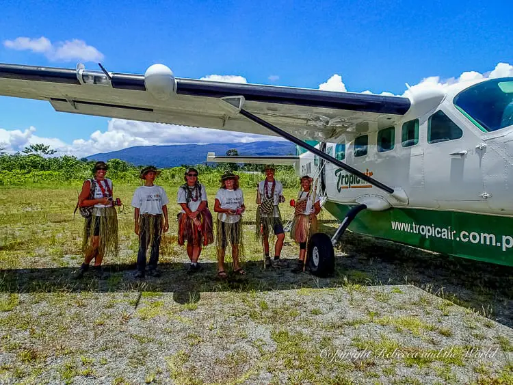 A group of Kokoda Track trekkers in traditional attire standing in front of a small plane on a grassy airstrip. The bright blue sky and mountainous backdrop indicate the remote and scenic location of their trek's endpoint.