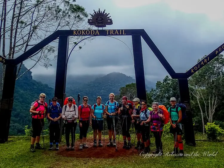A group of hikers standing under the Kokoda Trail archway, ready to begin their trek. They are dressed in outdoor gear and holding trekking poles. Behind them, lush mountains are partially obscured by mist.