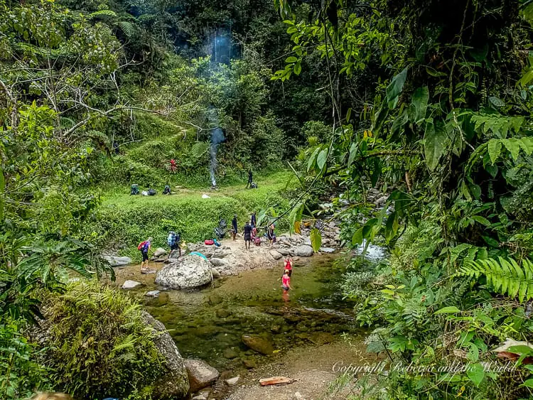 A group of hikers and guides resting by a riverbank, with some preparing to cross the water. The area is lush with greenery, and the river is calm and shallow. This guide shares what to expect when you walk the Kokoda Track in Papua New Guinea.