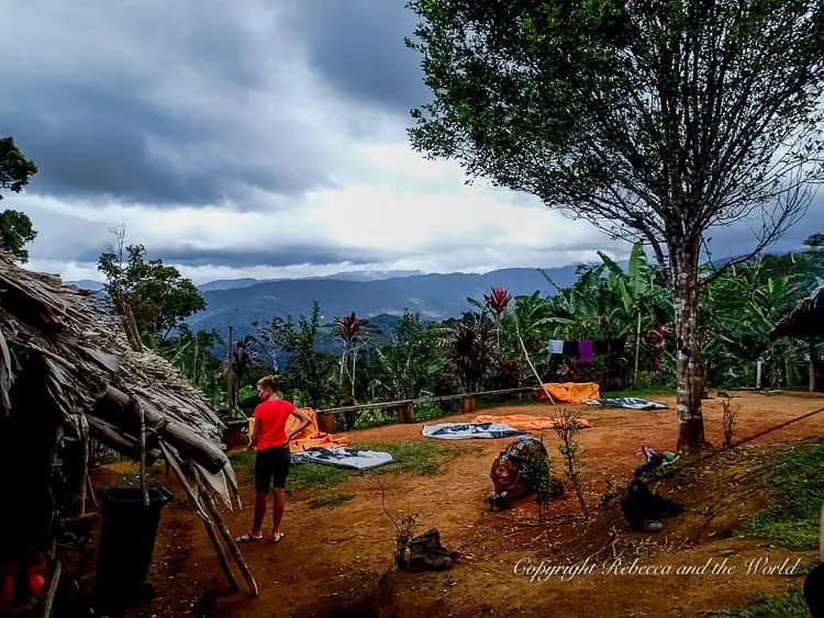 A campsite on the Kokoda Track with tents set up on a dirt clearing, surrounded by tropical plants and trees. A person in a red shirt is standing near a tree, while others are seated or tending to their gear.