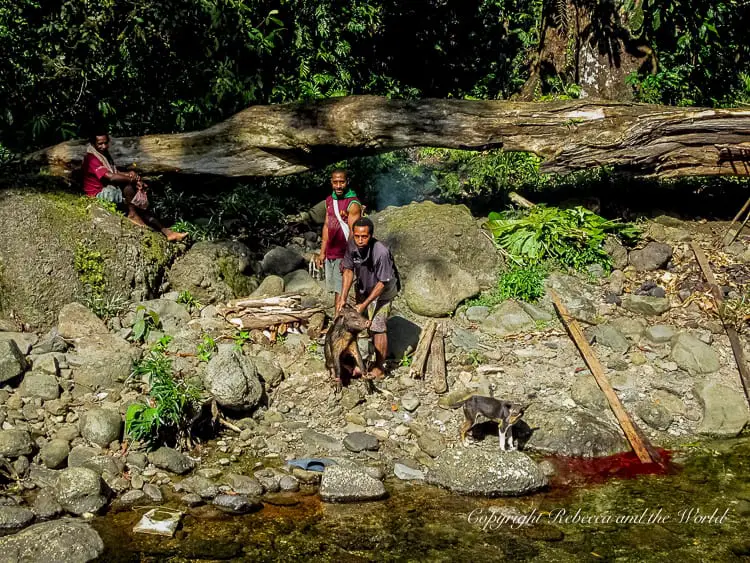 Three local men near a riverbank, preparing a fire and handling a dead pig. Two are standing, one is sitting on a rock, and a small dog is nearby. Large boulders and a fallen tree are in the background.