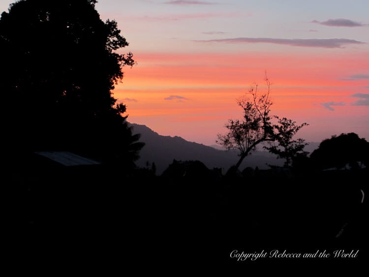 A serene sunset with a colorful sky of orange and pink hues. Silhouetted trees and distant mountains frame the scene, creating a peaceful atmosphere.