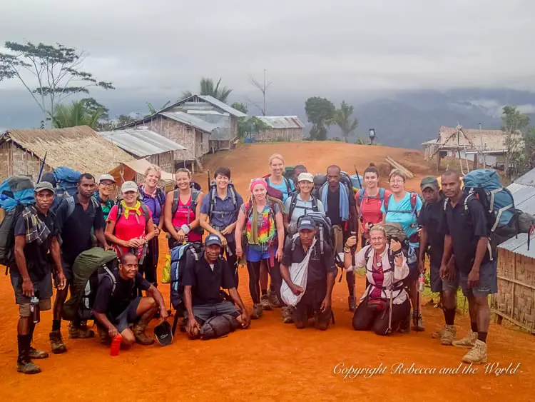 A group of Kokoda Track hikers and guides posing together in a village, with thatched-roof huts and mountains in the background. The trekkers have backpacks and appear ready for the journey ahead.