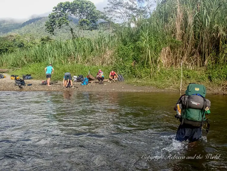 Hikers resting on a riverbank, with one person wading through the shallow water carrying a large backpack. The area is surrounded by tall grasses and mountains in the distance. The Kokoda Track includes a few river crossings.