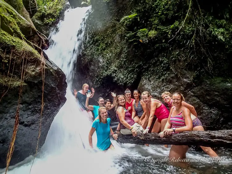 A group of women posing in front of a waterfall, sitting on rocks and smiling at the camera. They are wearing casual clothes and appear to be enjoying the natural setting, with water cascading down behind them.