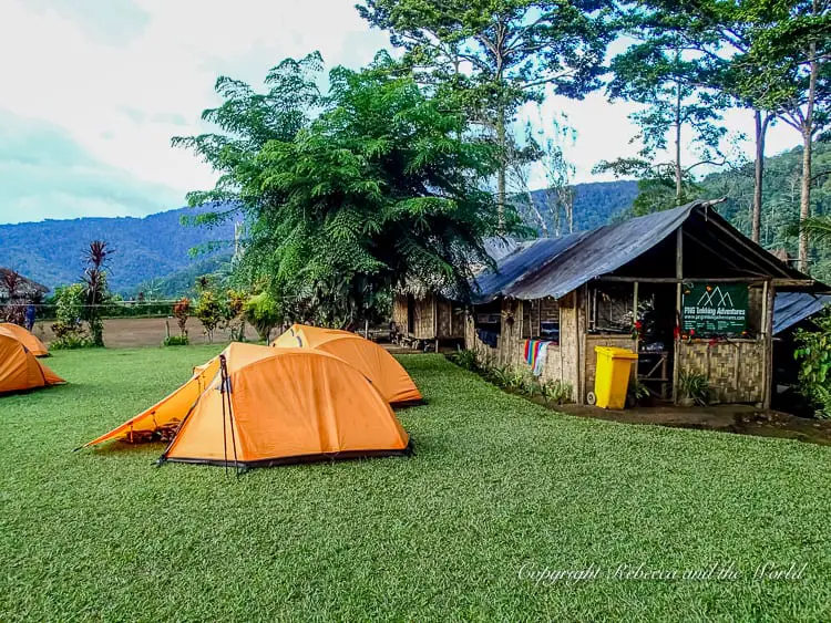 A campsite with several orange tents set up on a well-maintained grassy area. In the background, there are wooden buildings, trees, and mountains, creating a picturesque outdoor setting. Campsites along the Kokoda Track in Papua New Guinea are generally very picturesque.