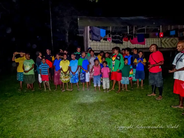 A group of local villagers in Papua New Guinea, including children and adults, gathered together at night on a grassy field. They are standing in a semi-circle, dressed in colourful clothing, and are singing.