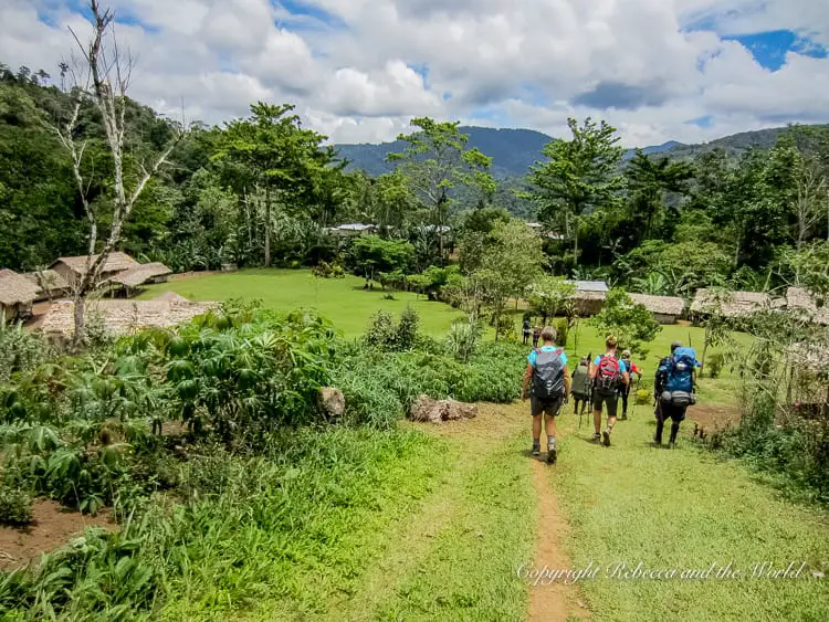 Five hikers walking down a grassy path towards a village with traditional thatched-roof huts. The path is surrounded by lush vegetation and trees, with mountains visible in the distance under a partly cloudy sky.