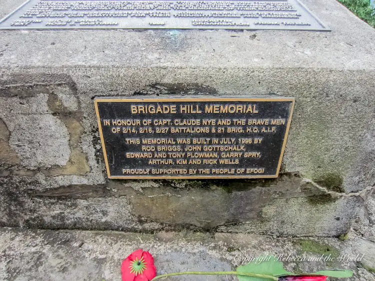A close-up of a memorial plaque on a stone monument, reading "Brigade Hill Memorial" and honoring Capt. Claude Nye and the brave men of various battalions. A single red flower lies at the base of the plaque.