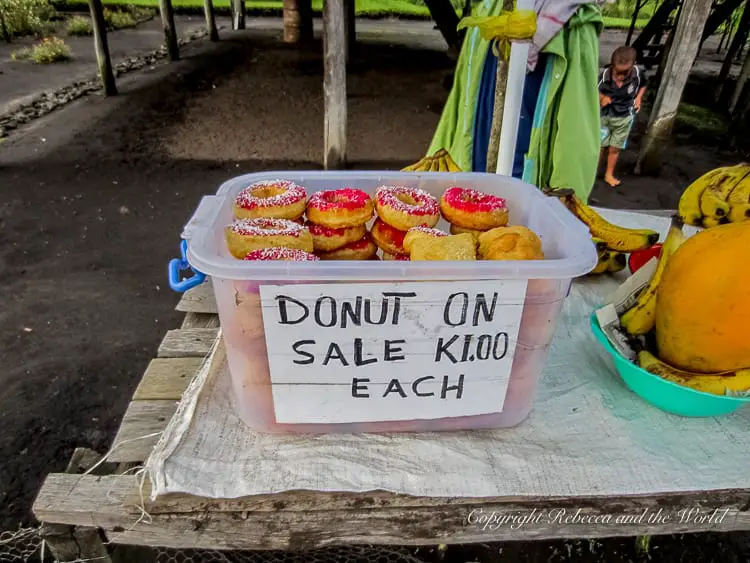 A plastic container filled with donuts, some topped with pink icing and sprinkles, displayed on a table for sale. A handwritten sign indicates the price of "K1.00 each." Fresh fruit is also visible nearby.