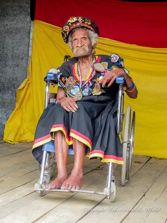 An elderly man sitting in a wheelchair, dressed in a traditional outfit adorned with various medals and decorations. A colourful backdrop of yellow and red fabric is behind him.This is Ovuru Ndiki, one of the last surviving "Fuzzy Wuzzy Angels".