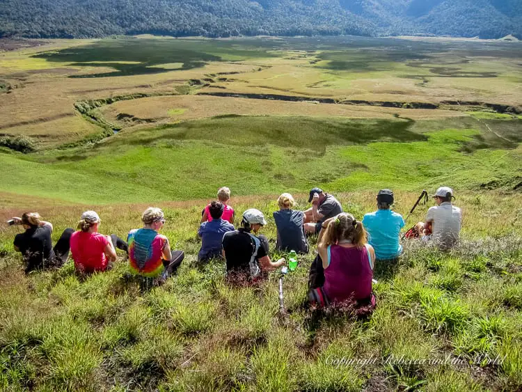 A group of hikers sitting on a grassy hill, taking a break and enjoying the expansive view of the green valley and mountains below. They are facing away from the camera, resting and chatting.