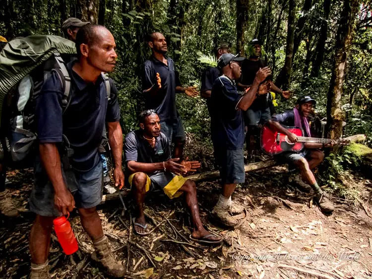 Local guides and porters singing and playing a guitar in a jungle setting. They are dressed in dark clothing, and some are clapping or gesturing as they perform, creating a lively and communal atmosphere.