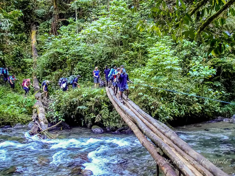 Hikers crossing a river on a makeshift bridge made of logs, with a rope for support. The surrounding area is densely forested, and the river below is fast-flowing. The Kokoda Track in Papua New Guinea includes several river crossings.