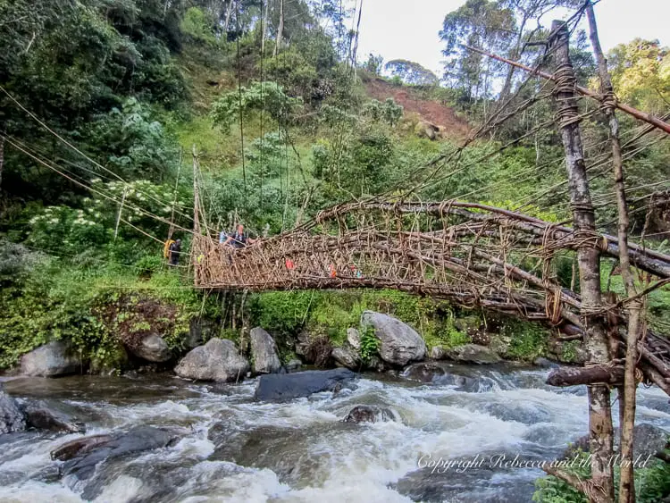 A rustic suspension bridge on the Kokoda Track made of woven vines and branches spans across a fast-flowing river. The bridge is surrounded by dense greenery, and a few people are visible on it, walking carefully.
