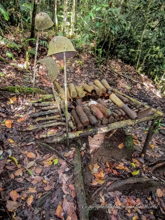A display of old, rusty artillery shells and helmets from World War II, arranged on a wooden frame in a jungle setting. The items are heavily corroded, showing their age and historical significance.