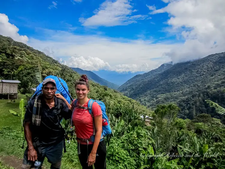 A man and a woman (the author of this article and her Kokoda Track porter/guide), smiling and posing for a photo against a backdrop of lush mountains and blue sky. They are wearing backpacks and trekking gear, indicating they are on a hiking adventure.