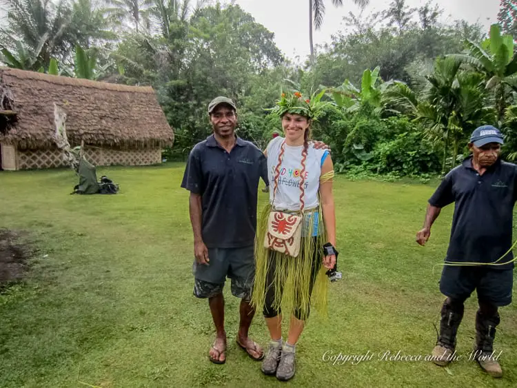 A female trekker - the author of this article - wearing a traditional grass skirt and flower crown standing next to a local guide in a dark blue shirt. They are posing for a photo on a grassy area with traditional huts and dense vegetation in the background.