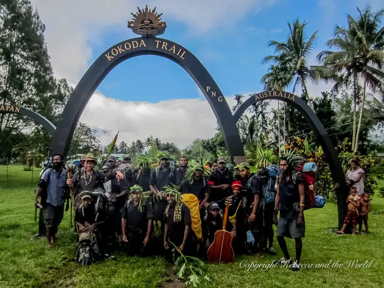 A group of trekkers and local guides standing under the archway marking the end of the Kokoda Trail. The arch is inscribed with "Kokoda Trail" and the flags of Papua New Guinea and Australia. The group looks happy and ready to end their journey.
