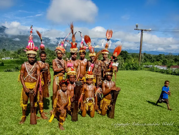A group of local performers, including men and boys, dressed in traditional attire with body paint, feathers, and shells. They are standing on a grassy field with mountains and blue sky in the background, showcasing the cultural heritage of the area. They performed on Kokoda Day at the end of the Kokoda Track.