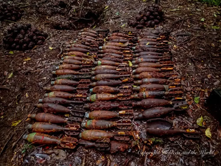 Rows of rusty, old mortar shells and other unexploded ordnance from World War II, arranged on the ground in the jungle. The items are heavily corroded, indicating their age and exposure to the elements.