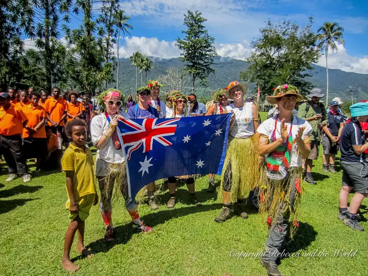 A group of Kokoda Track trekkers, some wearing grass skirts and flower crowns, holding an Australian flag and posing with local villagers. The scene is festive, with other people in the background and palm trees around, highlighting a moment of celebration.