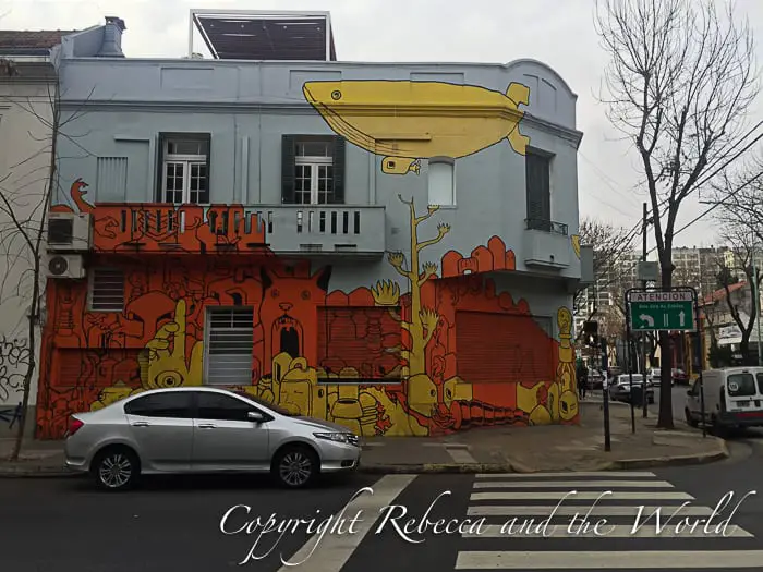 A car is parked in front of a grey building in Buenos Aires, Argentina. The building is covered in coloured street art.