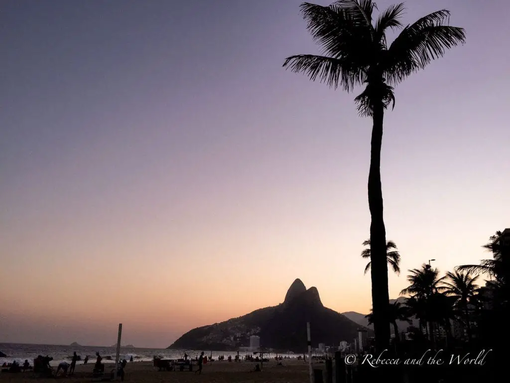 A silhouette of palm trees against a sunset sky, with the outline of a mountain in the distance. People are visible on a beach in the foreground. A visit to Rio de Janeiro in Brazil should be on everyone's travel bucket list.