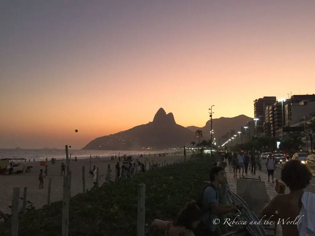 Sunset at Ipanema Beach in Rio de Janeiro with silhouettes of people and mountains in the background. The sky gradients from pink to blue, and a promenade on the right is lined with street lamps and buildings.
