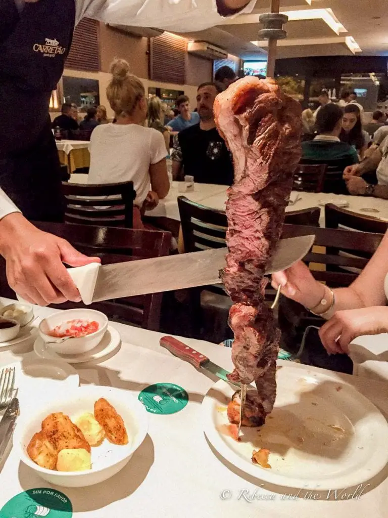 A server in a restaurant uniform holding a large skewer of roasted meat, ready to slice it onto a diner's plate. Other diners and tables are visible in the background. Brazilians love their beef and there's no better place to try it than at a churrascaria, where servers carve off slices off meat for you at the table.