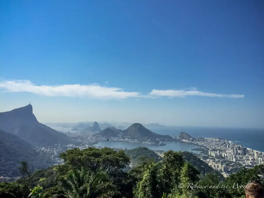 A panoramic view overlooking Rio de Janeiro from a high vantage point. Lush greenery in the foreground, with the city spread out towards the water and mountains in the distance. There are great panoramas of Rio de Janeiro from the world's largest urban rainforest, Parque Nacional da Tujica.