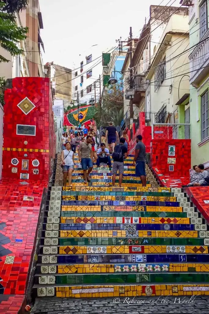 A stairway with colorful mosaic tiles, crowded with people. Red patterned tiles adorn the sides, and the stairs are in the center, leading up between two buildings. Escaderia Selaron is a popular Rio de Janeiro tourist attraction - it's great spot for photos.