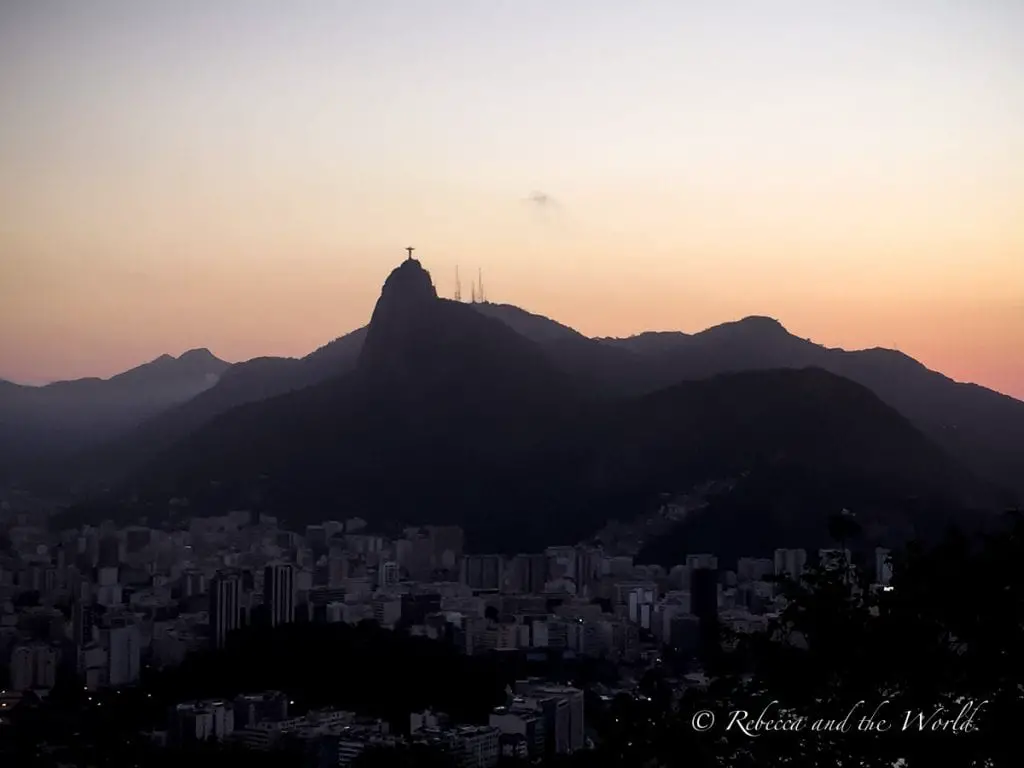 A twilight view of Rio de Janeiro with a mountain silhouette. The sky is in shades of purple and orange, with city lights beginning to twinkle. The view from Sugarloaf Mountain in Rio de Janeiro is a great place to watch the sunset.